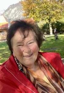 Das lebensfrohe Porträt einer älteren Frau mit braunem Haar und roter Jacke im herbstlichen Park – Ein Beispiel für gelebte Würde, Geborgenheit und das Lösen emotionaler Blockaden durch professionelle Aromatherapie, basierend auf der Expertise von Duftgenuss Barbara Tausch als SGZ-Dozentin und STV-PDL im Palliativcarehaus 