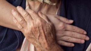 Eine behutsame, tröstende Handberührung, bei der eine jüngere Hand die gealterte Hand einer Seniorin im Bett hält – schmerzlindernde Aromapflege am Lebensende zur Linderung von Ängsten und Stress bei Duftgenuss.