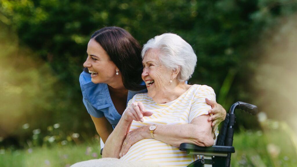 Eine junge Pflegekraft im blauen Kasack beugt sich im sonnigen Garten liebevoll zu einer älteren, glücklich lachenden Seniorin im Rollstuhl – professionelle Aromatherapie in der Palliative Care zur Steigerung der Lebensqualität von Duftgenuss.